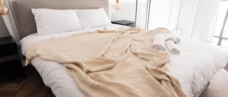 Una cama bien hecha con una manta color beige, almohadas blancas y toallas enrolladas al lado.