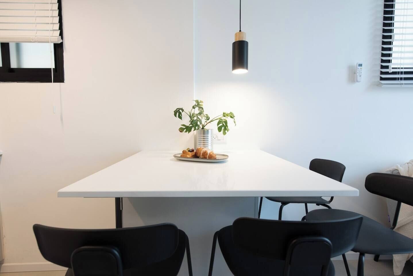 A modern dining area with a white table, black chairs, a hanging pendant light, and a potted plant.