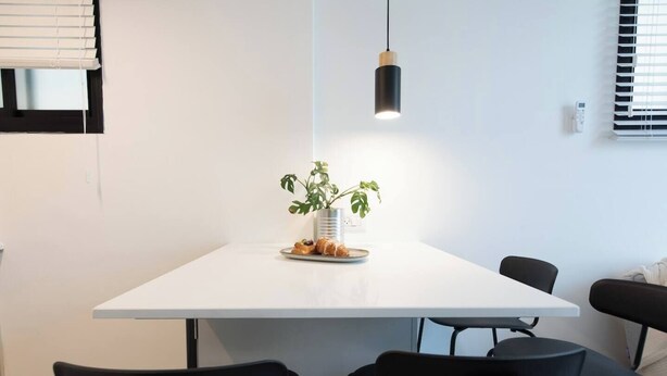 A modern dining area with a white table, black chairs, a hanging pendant light, and a potted plant. - Port city haifa-Urbanic Loft heart of haifa Dwntwn (Haifa)