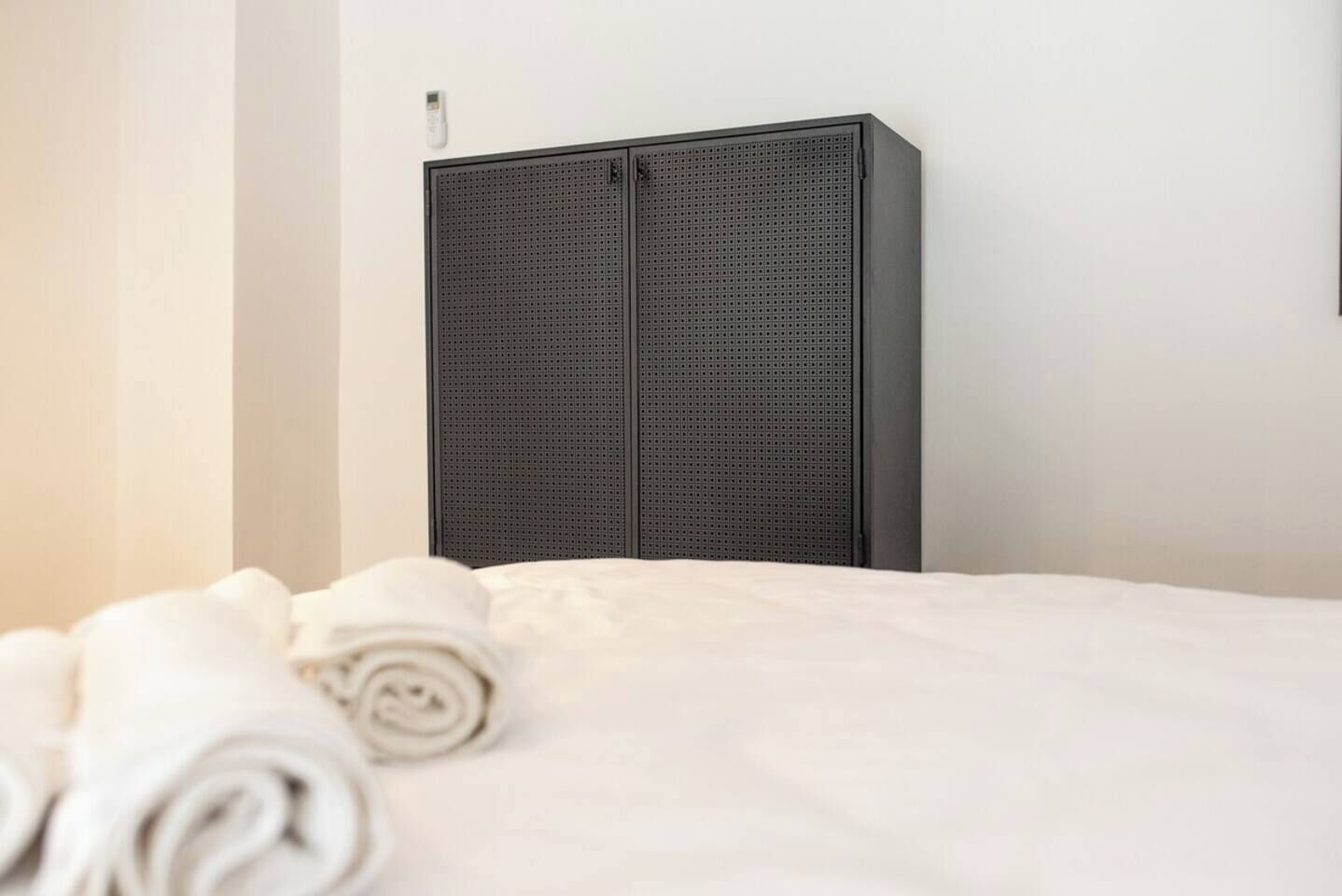 Una cama bien hecha con una toalla blanca enrollada, un armario oscuro con un pequeño dispositivo encima, y una pared blanca lisa.