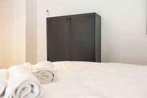 Ein ordentlich bezogenes Bett mit einem aufgerollten weißen Handtuch, ein dunkel gefärbter Kleiderschrank mit einem kleinen Gerät darauf und eine schlichte weiße Wand.