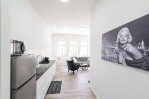 A modern kitchen with a refrigerator, microwave, and sink. There is a dining area with a table and chairs, and a large painting on the wall. - Port city haifa authentic 2bdrmsblcny germancolony (Haifa)