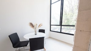 Ein Zimmer mit einem runden Tisch, zwei schwarzen Stühlen, einer Vase mit getrockneten Pflanzen und einem großen Fenster mit Blick auf Bäume.