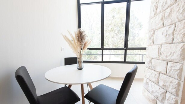 Ein Zimmer mit einem runden weißen Tisch, zwei schwarzen Stühlen, einer Vase mit Trockenpflanzen und einem großen Fenster mit Blick auf Bäume.