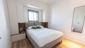 Un dormitorio con cama, mesita de noche, armario empotrado, ventana y espejo en la pared.