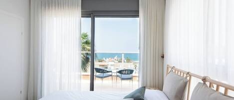 A hotel room with a bed, two pillows, a balcony with a table and chairs, and a view of the sea.