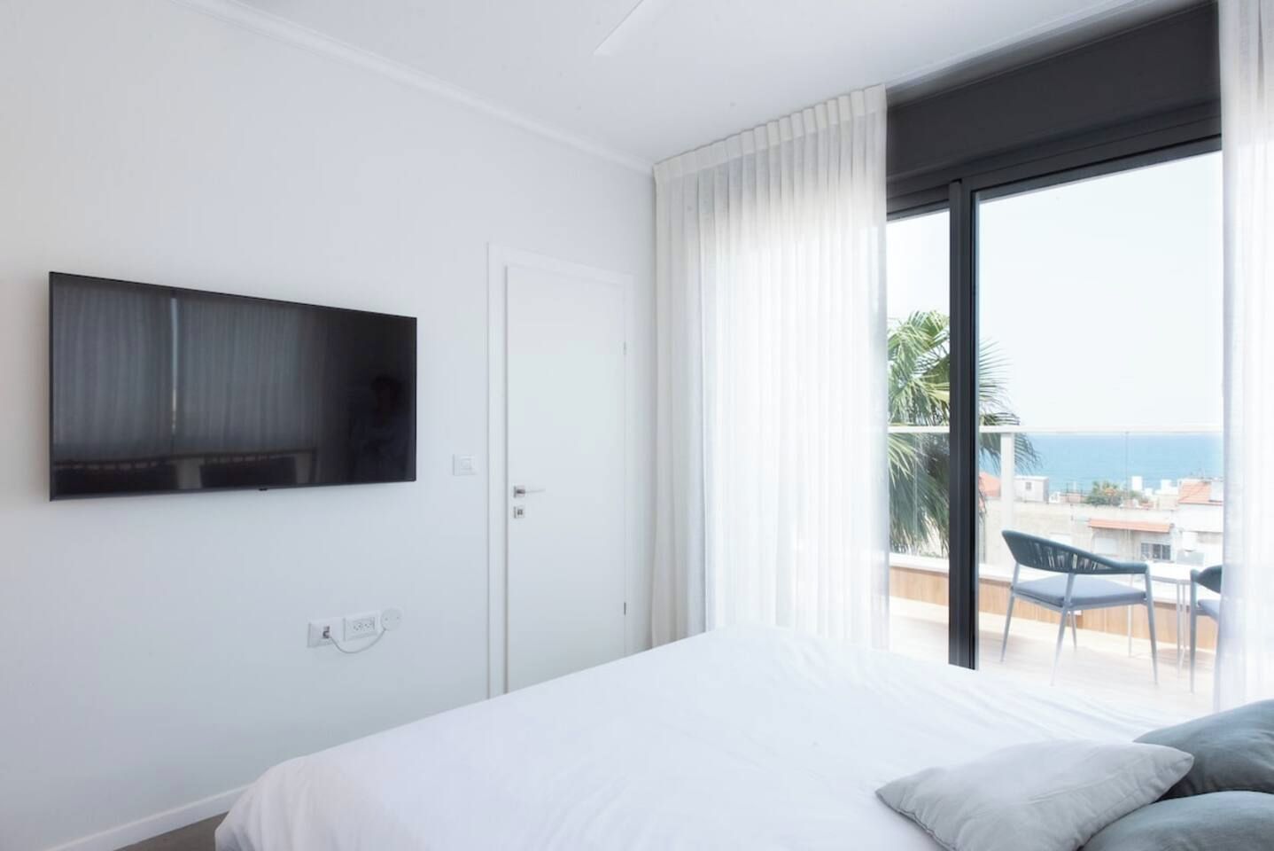 A bedroom with a bed, a TV mounted on the wall, a door, and a balcony with a view of the sea.