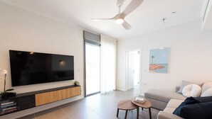 Ein modernes Wohnzimmer mit einem Flachbildfernseher, einem Deckenventilator, einer Couch, einem Couchtisch und einer Tür zu einem weiteren Raum.