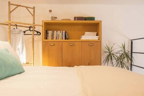 Een slaapkamer met een bed, een houten garderobe met boeken en handdoeken, een kledingrek en een potplant.