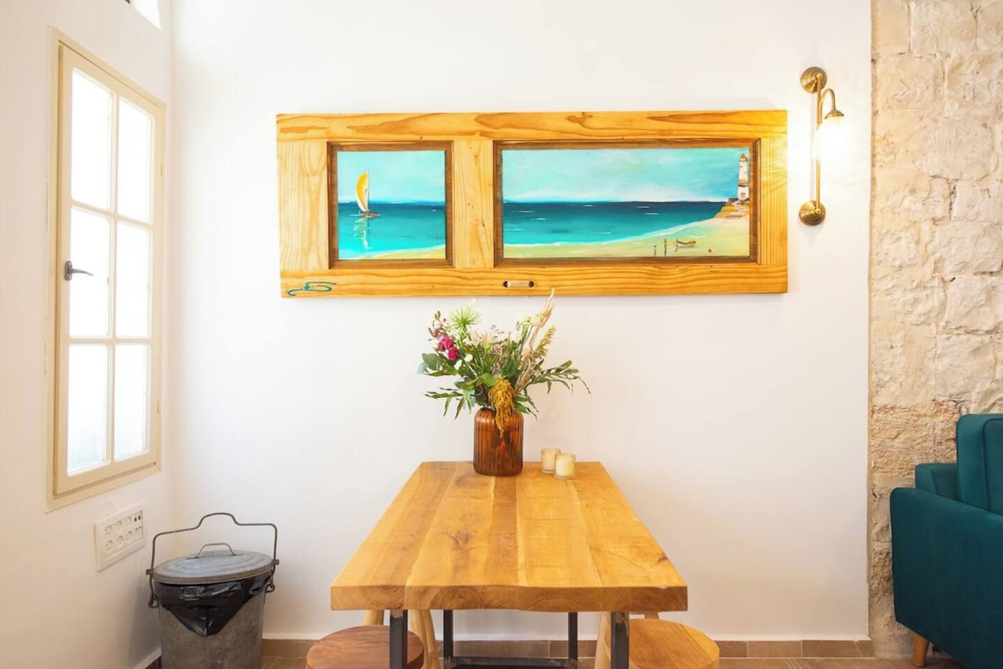 A room with a wooden table, a vase with flowers, a trash bin, and a wall-mounted picture of a beach scene.