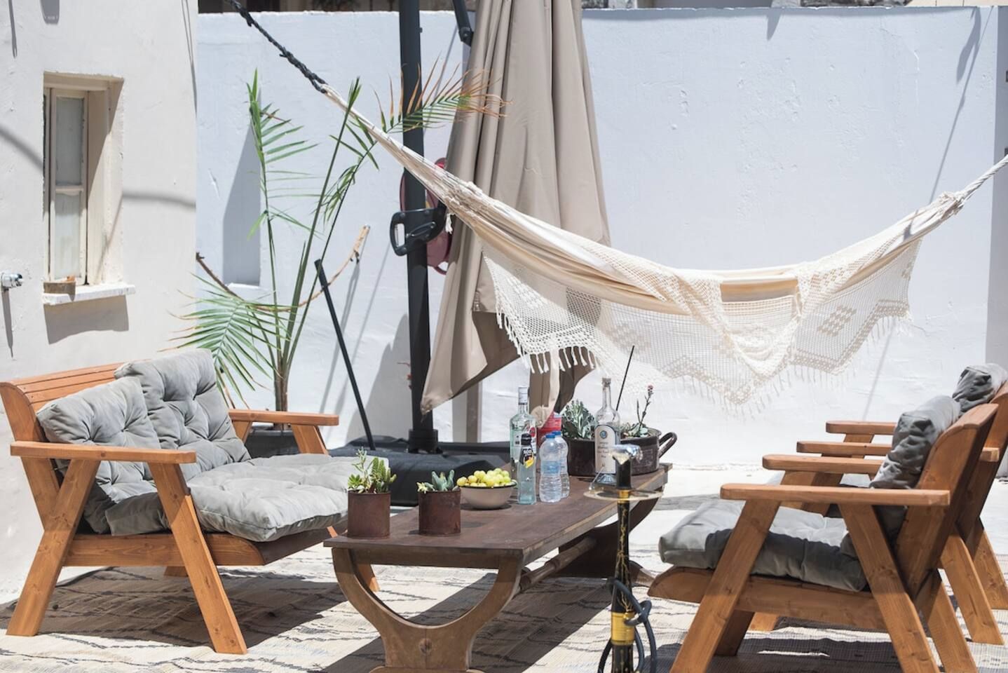 En hængekøje spændt ud mellem to træstole med puder, et lille bord med drikkevarer og planter, og en potteplante ved siden af stolene.