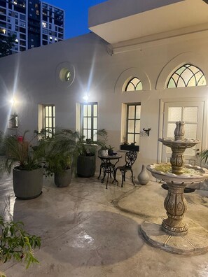 Un espace extérieur aménagé en patio, avec des plantes en pot, une table et des chaises, ainsi qu’une fontaine décorative.