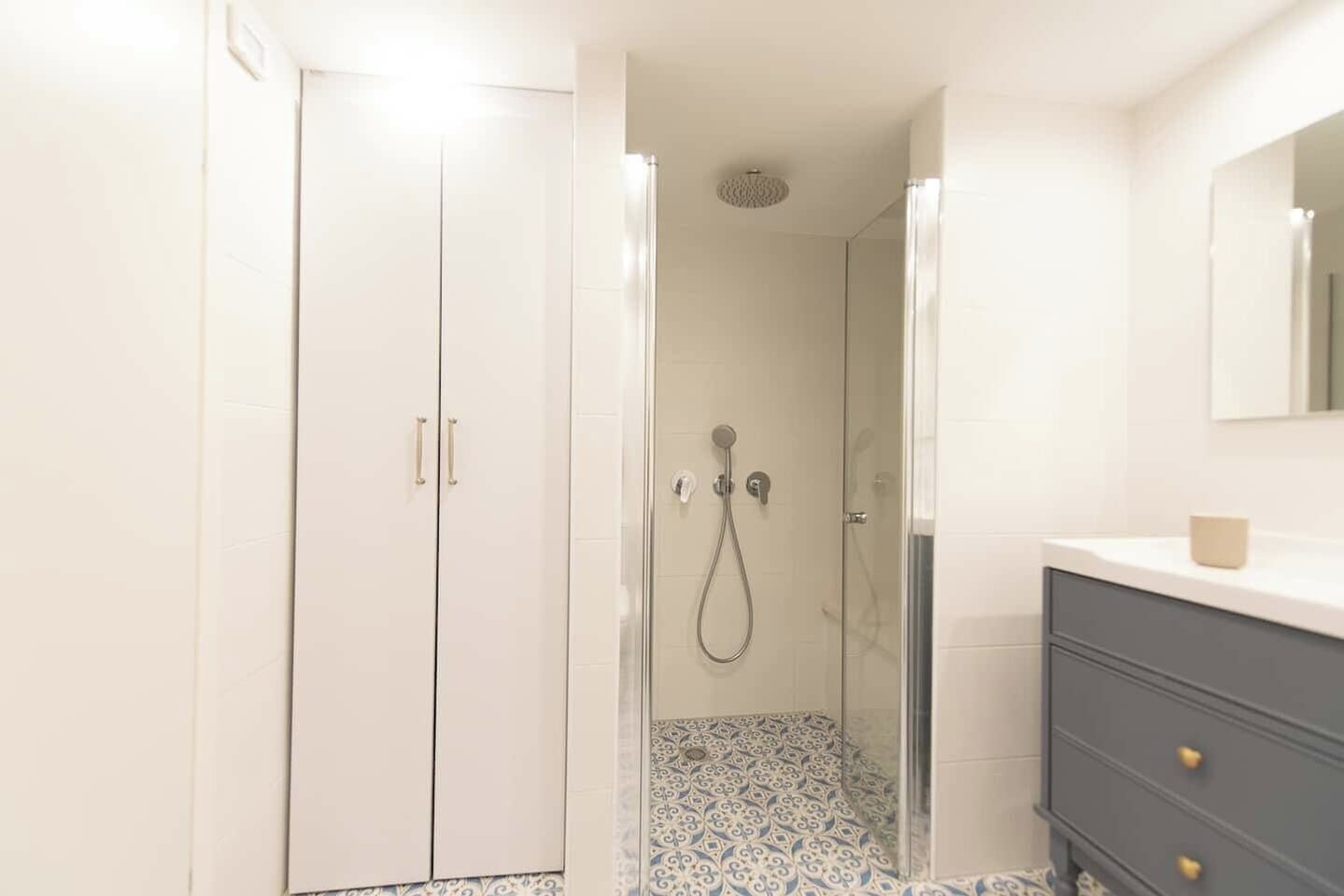A bathroom with a walk-in shower, a vanity with a mirror, and patterned floor tiles.