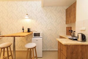 Eine kleine Küche mit Mikrowelle, einem kleinen Kühlschrank, Holzbarhocker und einer Arbeitsplatte mit Wasserkocher und weiteren Küchengeräten.