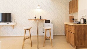 Uma cozinha compacta com micro-ondas, cafeteira e televisão sobre um suporte. Há dois bancos e uma pequena mesa com candeeiro.