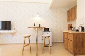 Eine kompakte Küche mit Mikrowelle, Kaffeemaschine und Fernseher auf einem Schrank. Zwei Hocker und ein kleiner Tisch mit Lampe stehen bereit.