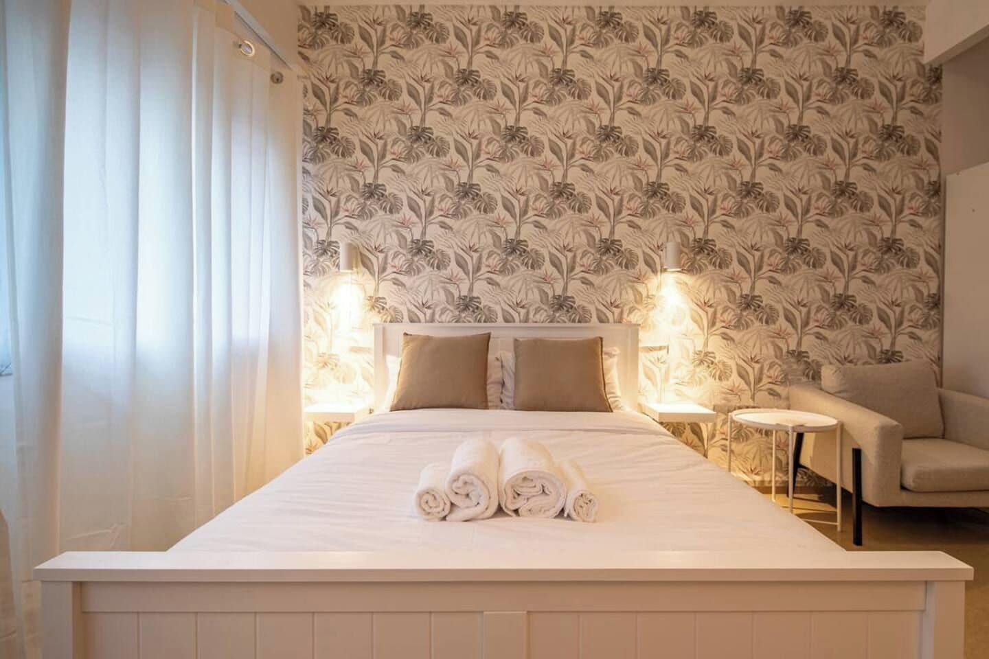 A hotel room with a large bed, two bedside lamps, a chair, and a floral wallpaper pattern.
