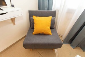 A grey sofa with a yellow pillow and a grey cushion in a room with a white desk and a computer monitor. - Downtown Haifa Studio 213, Near Port, AC & WiFi (Haifa)