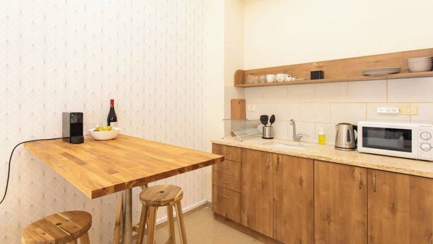 Uma cozinha compacta com uma mesa de madeira, banquetas, uma garrafa de vinho, cafeteira, chaleira e micro-ondas.