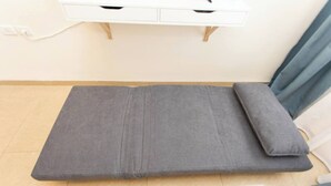 Um colchão cinzento sobre um chão de azulejos, com uma mesinha de cabeceira branca e uma cortina azul ao fundo.