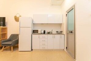 A compact kitchen with white cabinets, a microwave, and a refrigerator. There's a gray armchair and a wooden shelf with books. - Port city haifa - studio 214 (Haifa)
