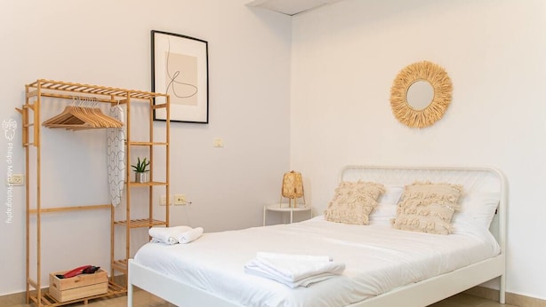 Ein ordentlich eingerichtetes Schlafzimmer mit einem weißen Bett, einem Holzschrank, einem kleinen Nachttisch und einem wandmontierten Spiegel.