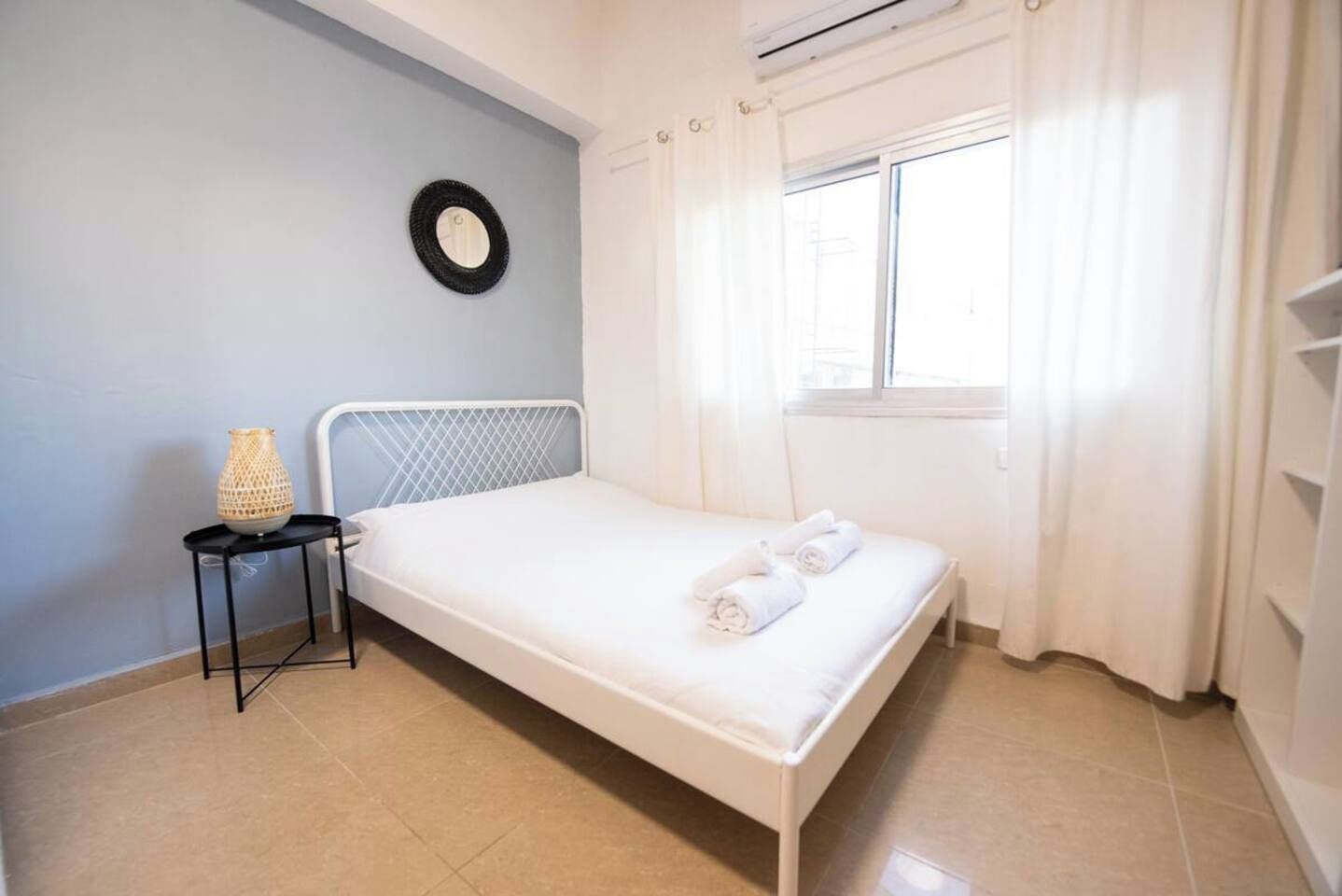 Une chambre avec un lit simple, une tête de lit en métal, une petite table avec une lampe, une fenêtre avec des rideaux et un miroir rond fixé au mur.