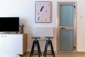 A modern room with a television on a wooden cabinet, two stools, a framed abstract painting, and a frosted glass door. - Port city haifa - Studio 218 (Haifa)