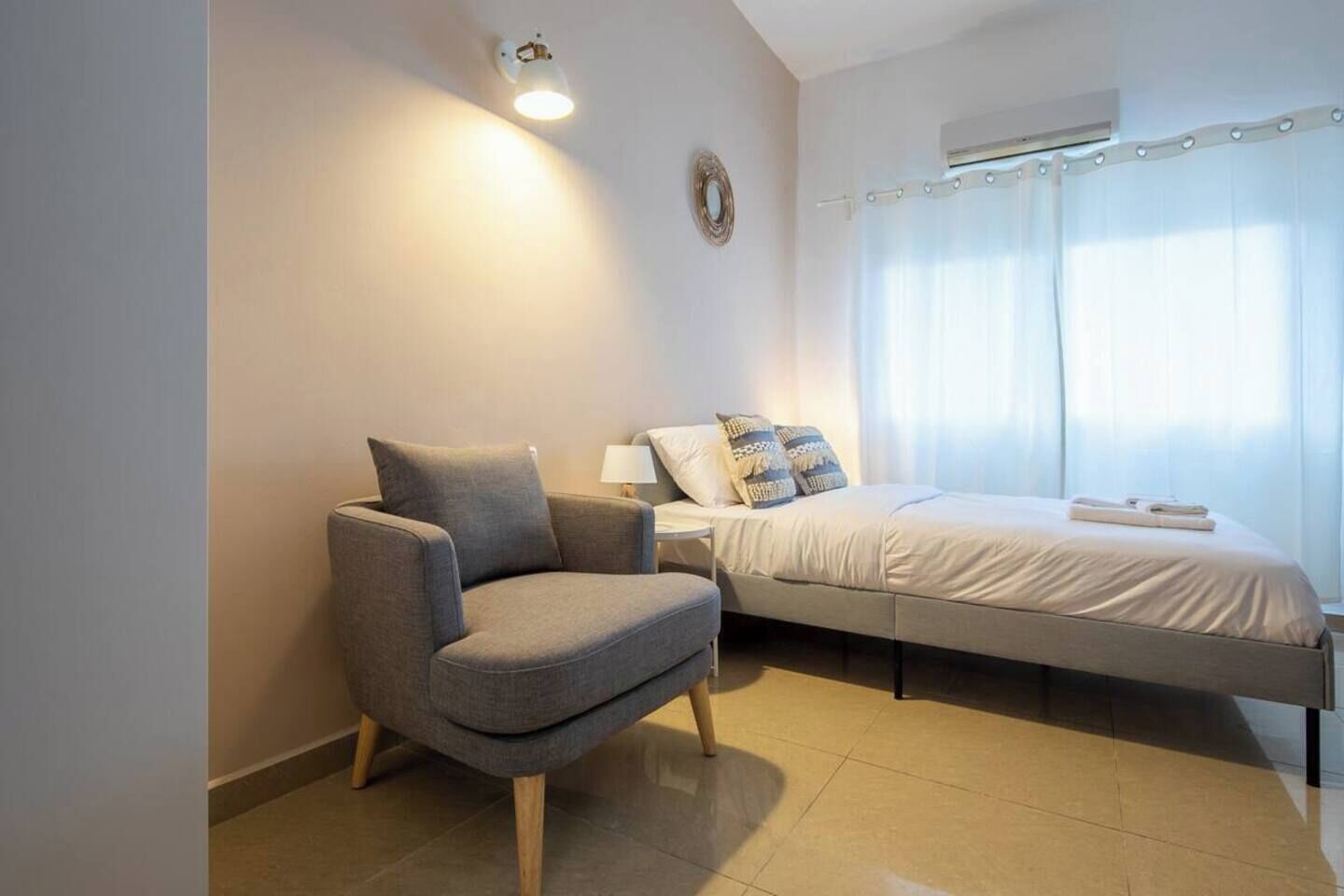 Un dormitorio moderno con una cama individual, un sillón gris, una mesita de noche con lámpara y una ventana con cortinas.