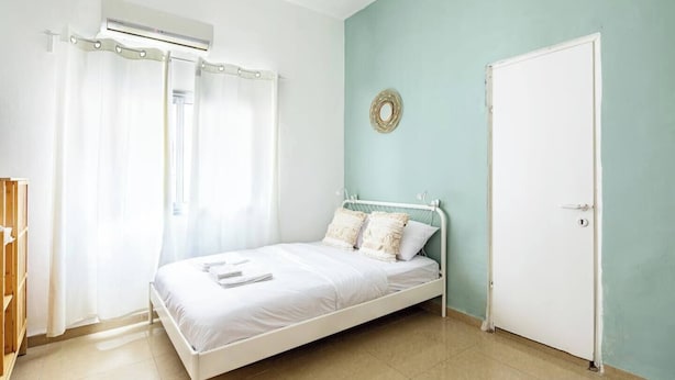 A bedroom with a bed, a white door, a window with curtains, and a wall-mounted air conditioning unit.