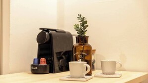 A coffee machine, a bottle with a plant, and two white cups on a wooden table.