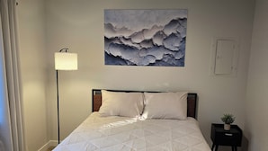 A neatly made bed with a quilted white duvet, a bedside lamp, a small potted plant, and a wall-mounted abstract painting.