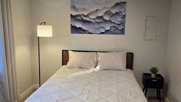 A neatly made bed with a quilted white duvet, a bedside lamp, a small potted plant, and a wall-mounted abstract painting.