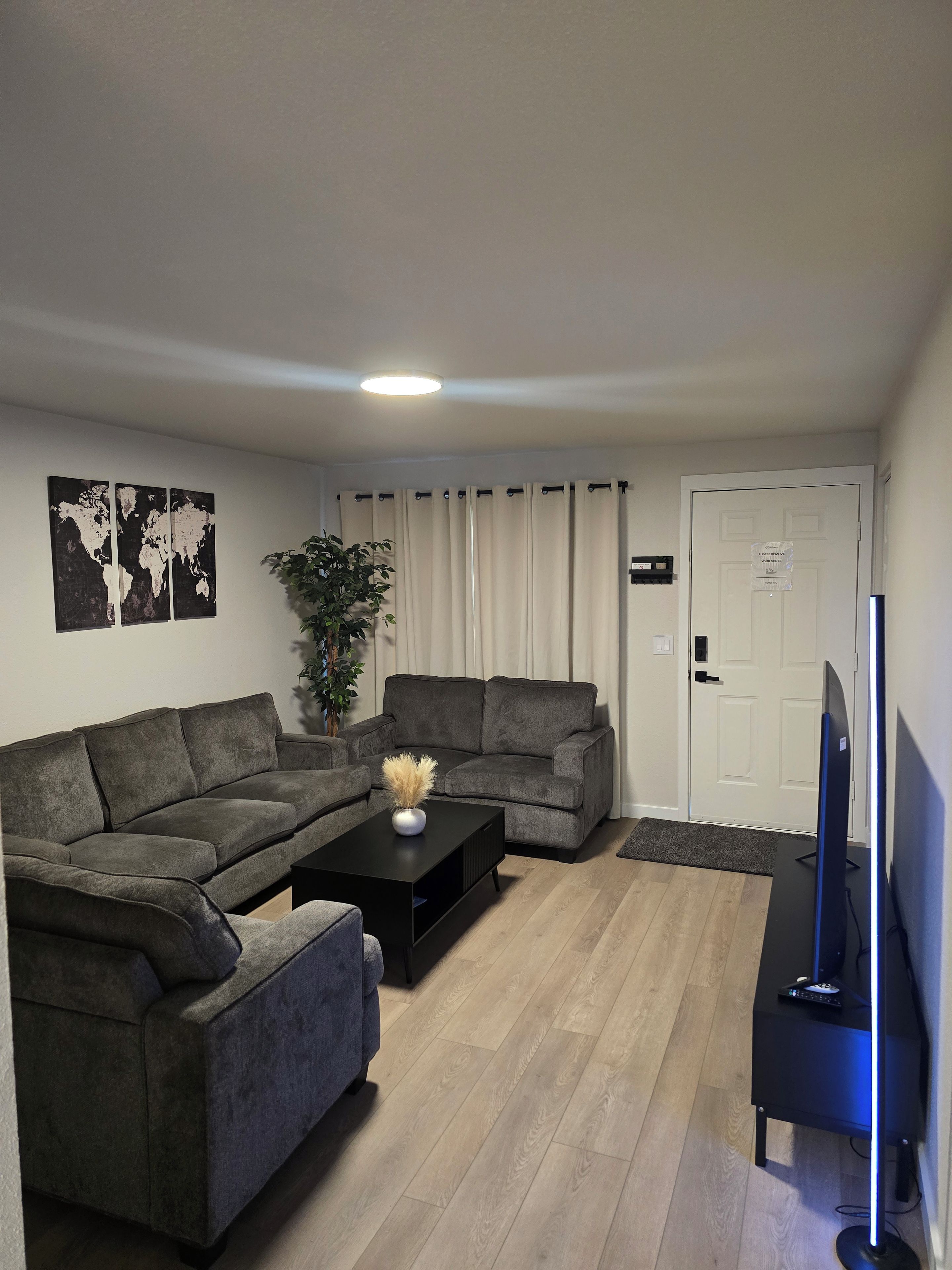 Una sala de estar con dos sofás grises, una mesa de centro negra, una planta, un mapa mundial en la pared, una puerta con un letrero y un mueble de TV con televisor.