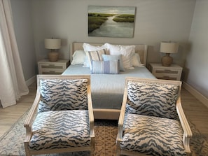 A bedroom with a bed, two bedside tables, two lamps, a painting on the wall, and two patterned armchairs.