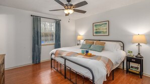 A neatly arranged bedroom with a bed, bedside lamps, a ceiling fan, a window with curtains, and a framed picture on the wall.