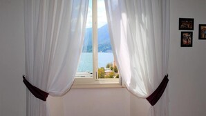 Una habitación con una ventana con cortinas blancas translúcidas y un lazo marrón oscuro en la parte inferior. Hay dos cuadros enmarcados en la pared a la derecha de la ventana.