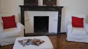 Una habitación con chimenea, dos sillones blancos con cojines rojos y una revista sobre un otoman pequeño.