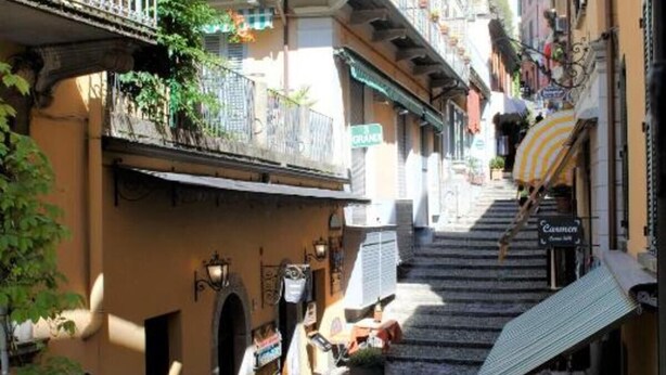 Una calle estrecha flanqueada por edificios, con una escalera que asciende y un pequeño espacio al aire libre con mesas y sillas.