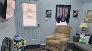 A living room with a beige armchair, a grey sofa, a standing fan, a wall-mounted air conditioner, and a door with blinds. - The Blue Goose, a charming house in Marshall, perfect for your getaway. (Marshall)