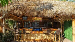Un área de bar con techo de paja, un letrero que dice "Aloha" y una barra con sillas.