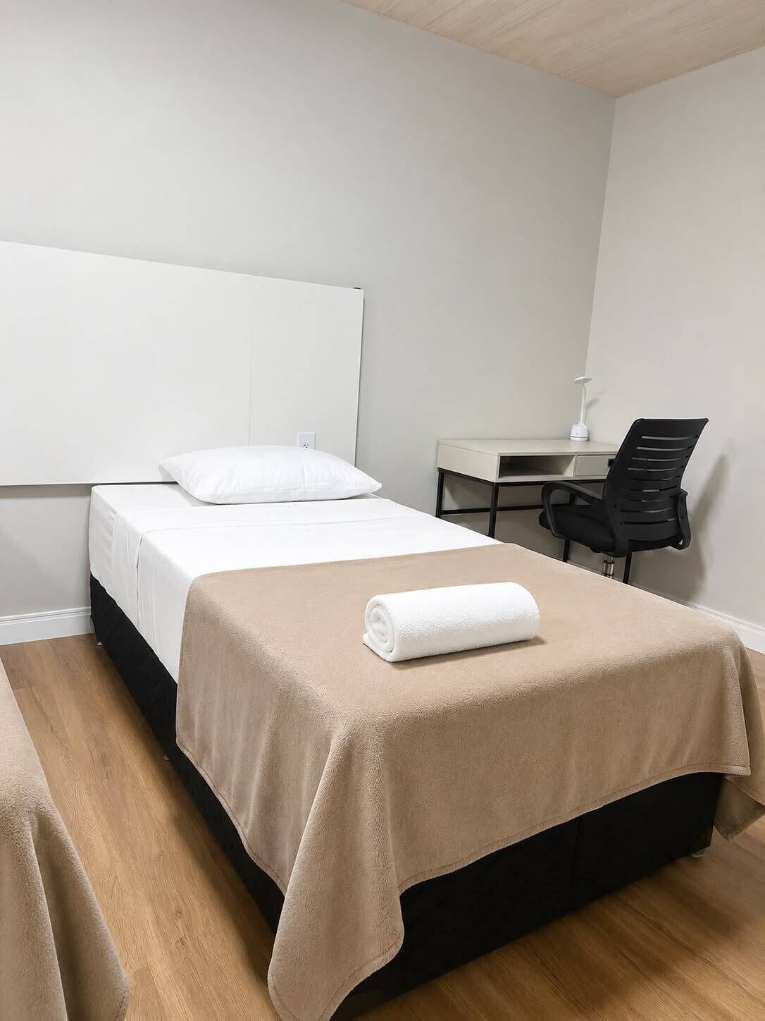 Una cama individual con cabecera blanca, un escritorio con silla y una toalla enrollada sobre la cama.