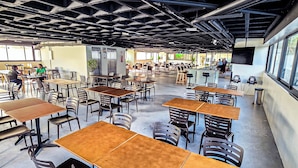 Un vaste espace intérieur doté de nombreuses tables et chaises, d’un plafond haut avec des poutres apparentes et de grandes fenêtres laissant entrer la lumière naturelle.