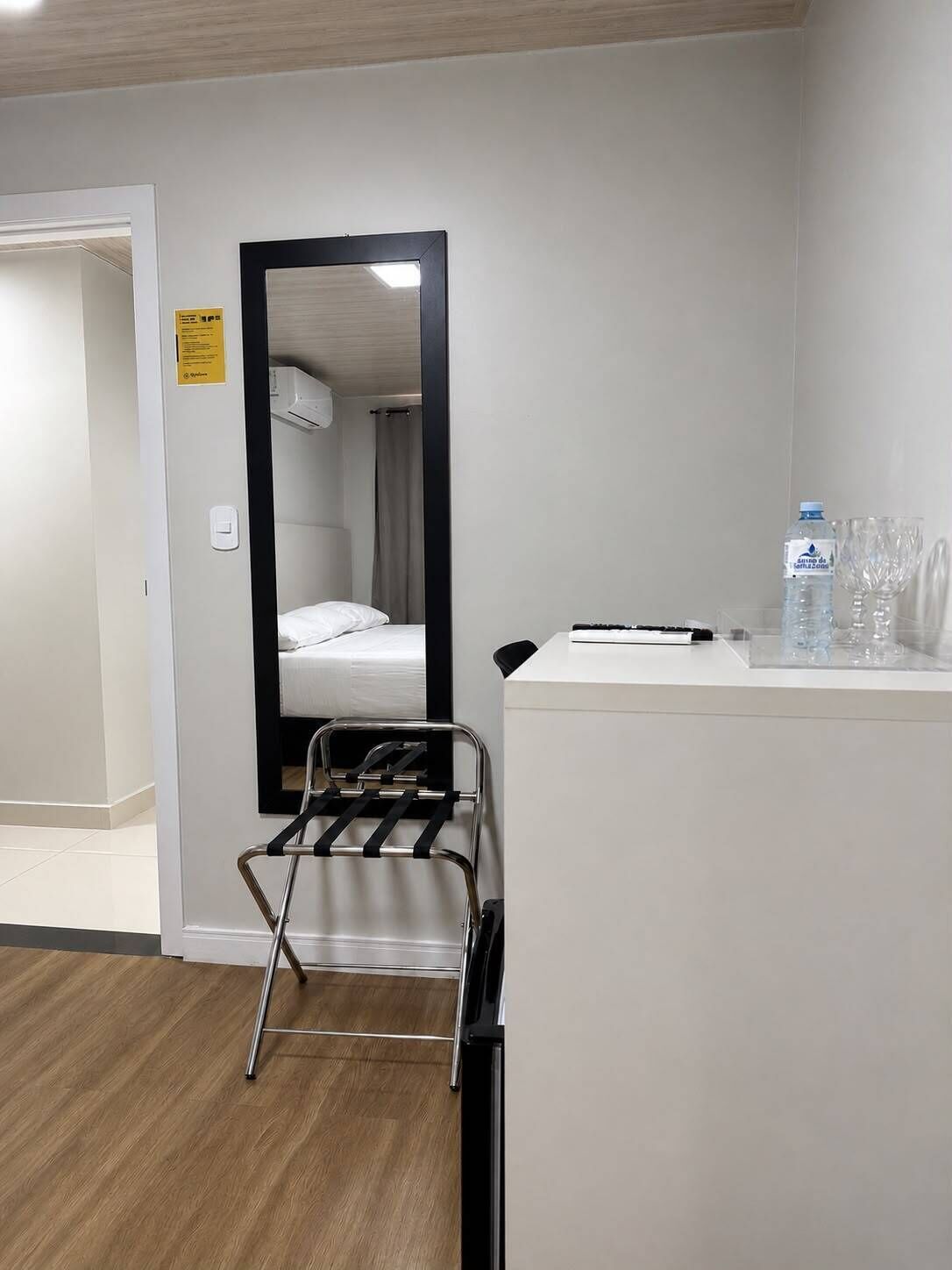 Habitación de hotel con cama, espejo, mesita con botella de agua y vasos, y silla con toallero.