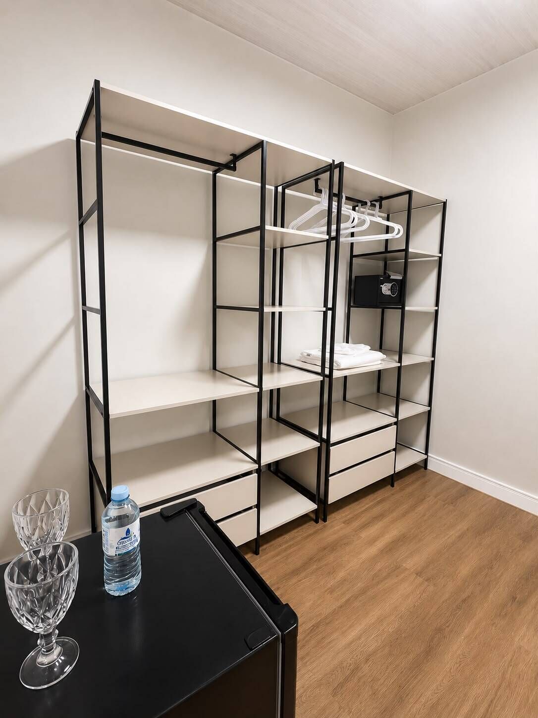 Een kamer met een zwarte metalen schapunit tegen een witte muur, een zwart tafeltje met een waterfles en een glas, en een houten vloer.