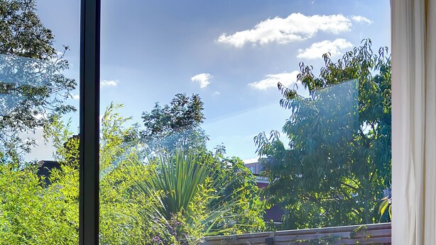 A sunlit patio with a metal chair and a cushioned bench, surrounded by lush greenery and a wooden fence. - Large, architect designed family home with garden close to tube in North London (London)