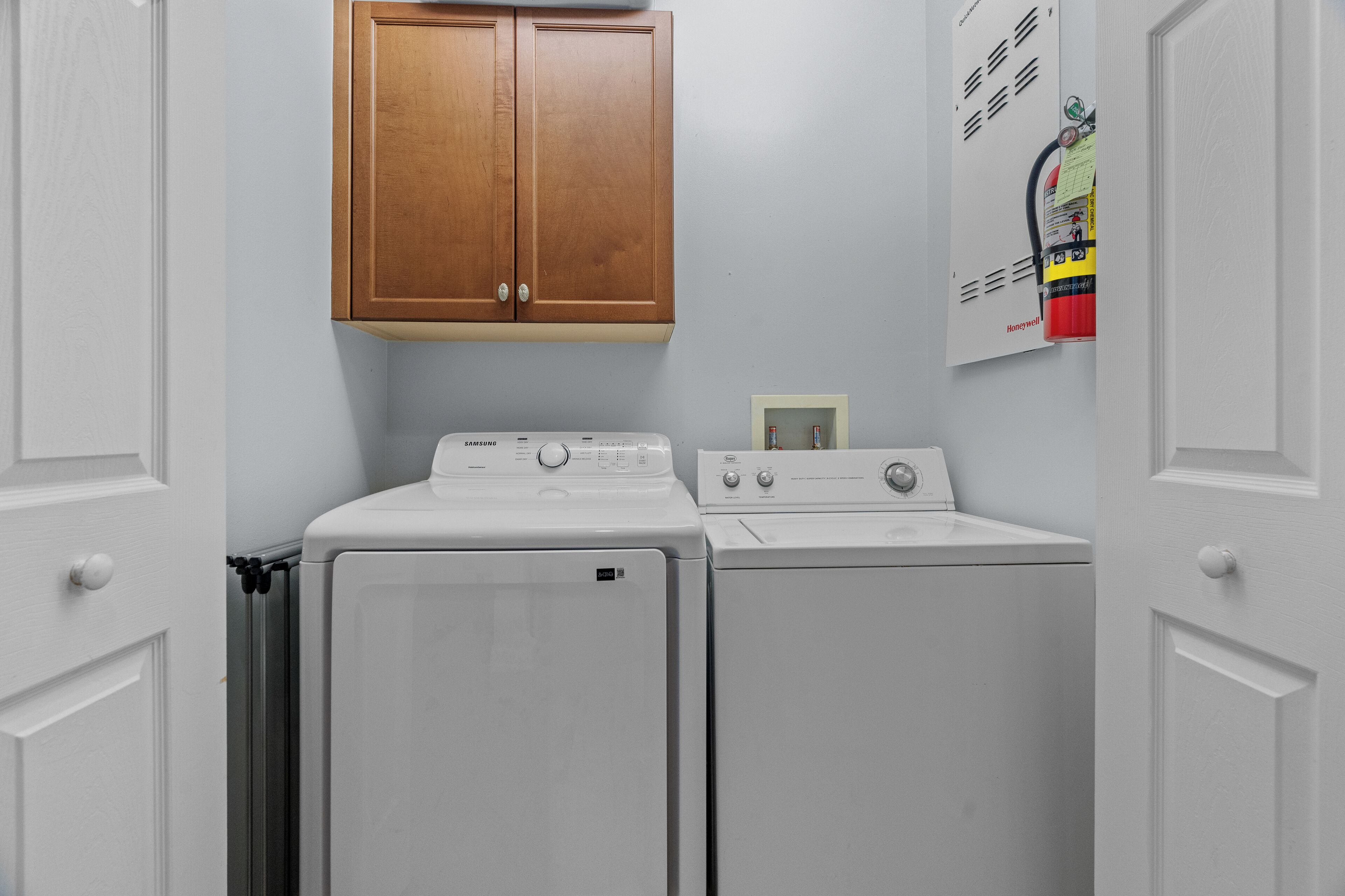 A laundry room with two white washers and dryers, wooden cabinets above, and a fire extinguisher on the wall.