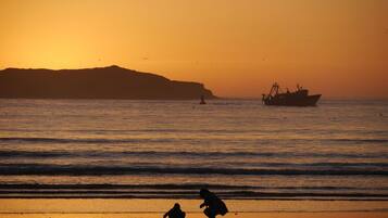 Une scène de plage au coucher du soleil, avec des silhouettes de personnes et un bateau de pêche sur l’eau.