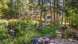 A stone pathway leading to a wooden deck with outdoor furniture, surrounded by lush greenery and a waterfall. - Luxury Vail Mountain Estate – Ski Access • Spacious • Elegant Mountain Retreat (Vail)