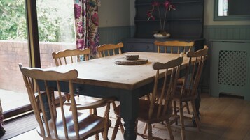 Una mesa de comedor con sillas, un mantel tejido y una cortina floral en una habitación con pared de ladrillo y ventana.